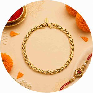 A gold-toned braided chain bracelet arranged in a circle on a warm beige surface with decorative orange flowers and a red-and-gold Rakhi thread visible at the lower right.
