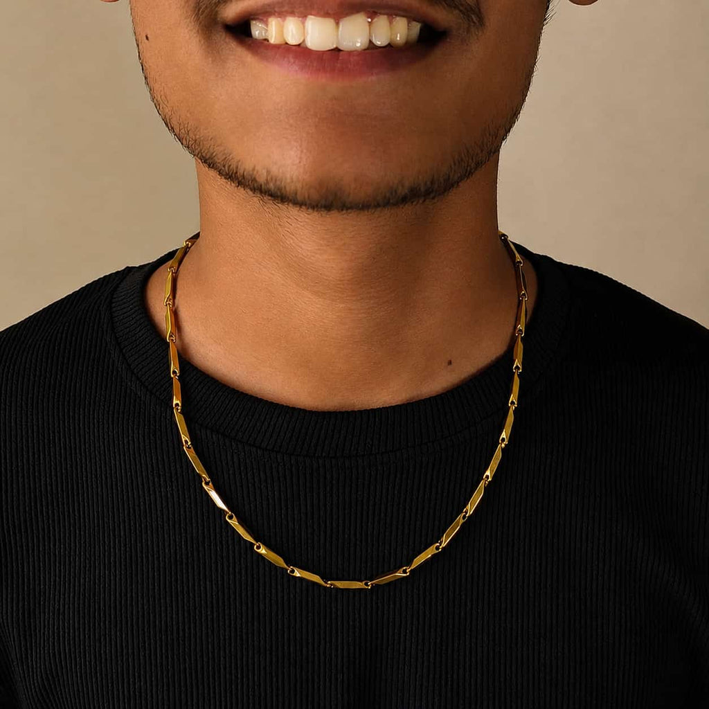 A close-up of a man wearing a gold chain necklace with elongated oval links, paired with a black ribbed t-shirt against a neutral beige background.