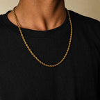 A close-up of a person wearing a gold rope chain necklace over a black ribbed crewneck shirt, set against a warm beige background.