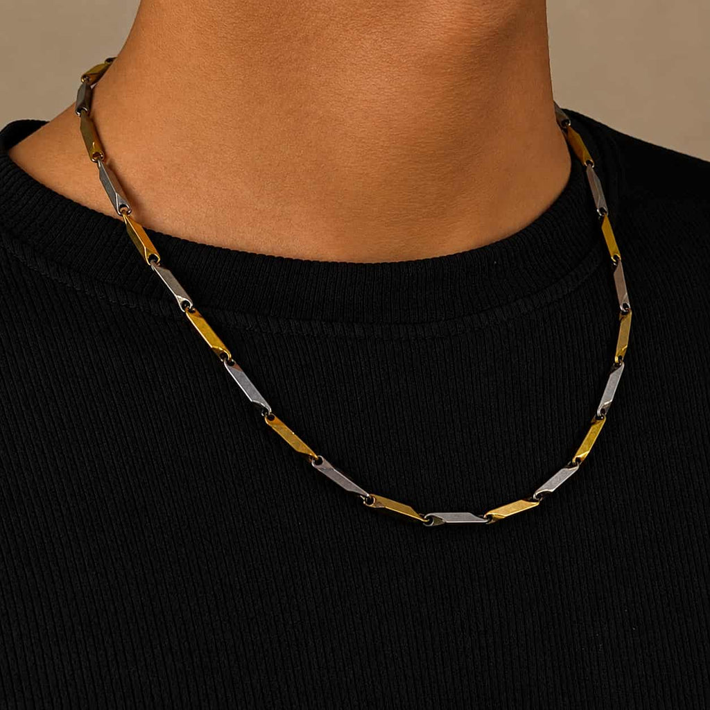 A close-up of a person wearing a two-tone metal link necklace with alternating gold and silver-colored oval links, paired with a black ribbed crewneck shirt.