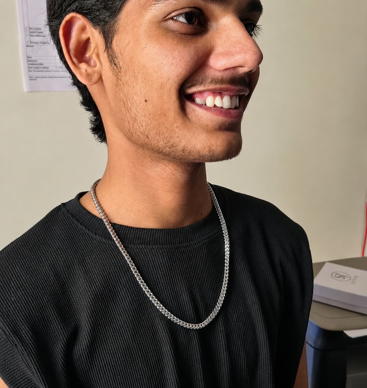 A smiling man wearing a silver chain necklace over a black ribbed t-shirt, photographed in an indoor setting.
