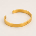 A polished gold-colored metal cuff bracelet with a minimalist design and a sliding clasp, displayed on a clean, light off-white background.