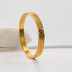 A gold-plated bangle bracelet with a textured pattern and an Om symbol engraved on the side, displayed on a white surface.