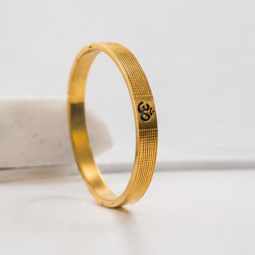 A gold-plated bangle bracelet with a textured pattern and an Om symbol engraved on the side, displayed on a white surface.