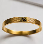 A gold-plated metal bangle bracelet with a textured honeycomb pattern and an Om symbol engraved on the front, displayed on a light gray surface.