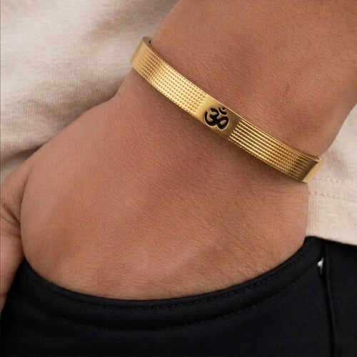 A gold-toned metal bracelet with a black Om symbol engraved on the front, shown worn on a person's wrist.