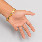 A gold-toned metal chain bracelet with textured and polished rectangular links, shown worn on a person's wrist with the hand palm facing forward against a light gray background.