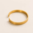 A gold-toned metal bangle bracelet with a textured geometric pattern on the outer band, shown on a plain light beige background.