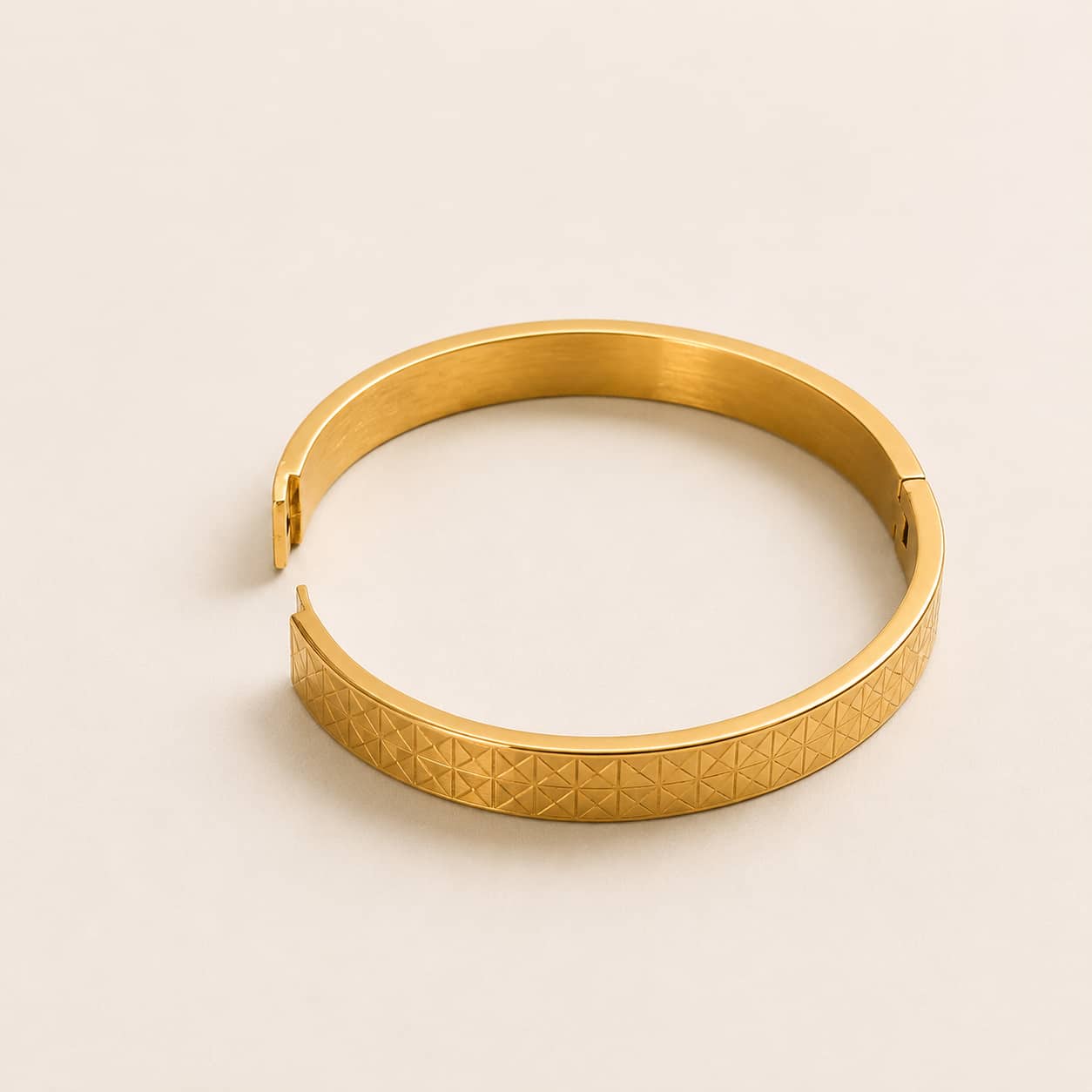A gold-toned metal bangle bracelet with a textured geometric pattern on the outer band, shown on a plain light beige background.
