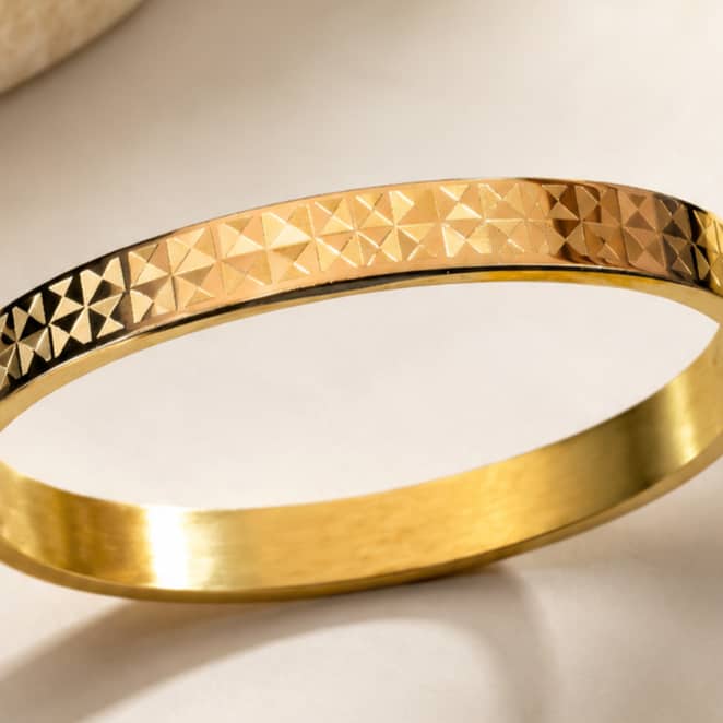 A gold bangle bracelet with a textured, geometric diamond-patterned outer band and a smooth, polished inner band, shown on a light beige background.