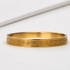 A gold bangle bracelet with a Greek key pattern engraved on the outer band, displayed on a clean white surface.