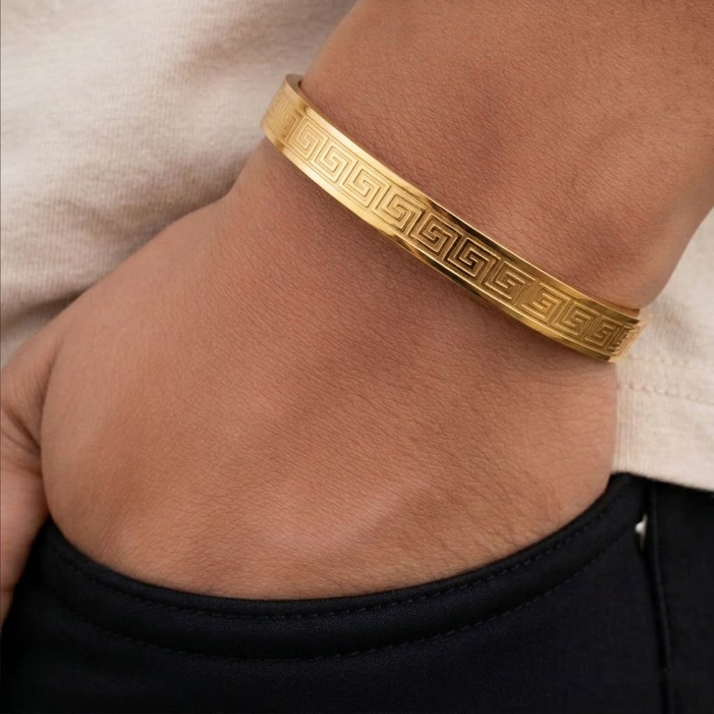 A gold bangle bracelet with a Greek key pattern, worn on a person's wrist.