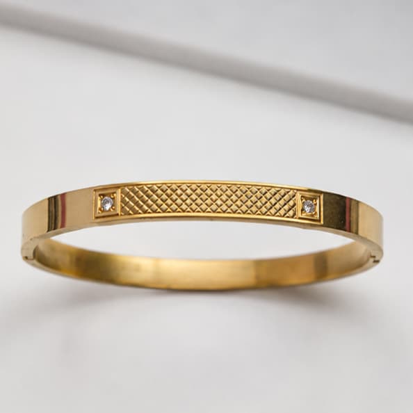 A gold bangle bracelet with a textured diamond-patterned center and two small clear gemstones on either side, shown on a light gray background.