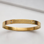A gold bangle bracelet with a textured diamond-patterned center and two small clear gemstones on either side, shown on a light gray background.