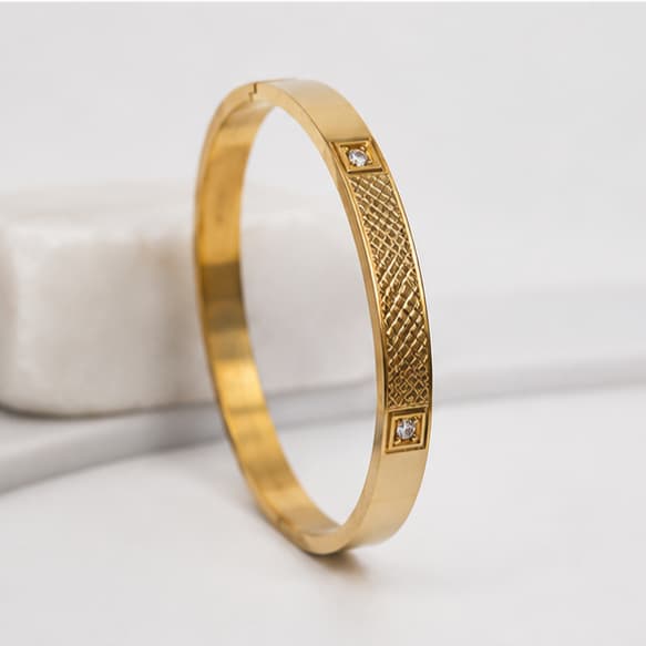A gold-plated bangle bracelet with a textured basketweave pattern and two small square crystal accents, displayed on a light gray surface.