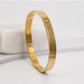 A gold-plated bangle bracelet with a textured basketweave pattern and two small square crystal accents, displayed on a light gray surface.