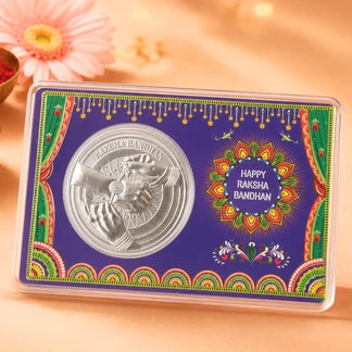 A silver Raksha Bandhan coin inside a clear plastic case, mounted on a decorative purple card with colorful traditional patterns and the text 'Happy Raksha Bandhan'.