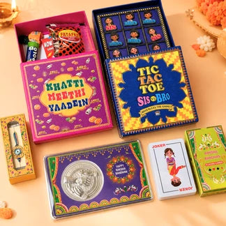 A collection of colorful Indian-themed gift boxes and cards arranged on a peach fabric background. Visible items include a pink 'Khatti Meethi Yaadein' box, a blue 'Tic Tac Toe Sis & Bro' box, a small 'Fatafi' box, a 'Joker' playing card, and other decorative gift packaging.