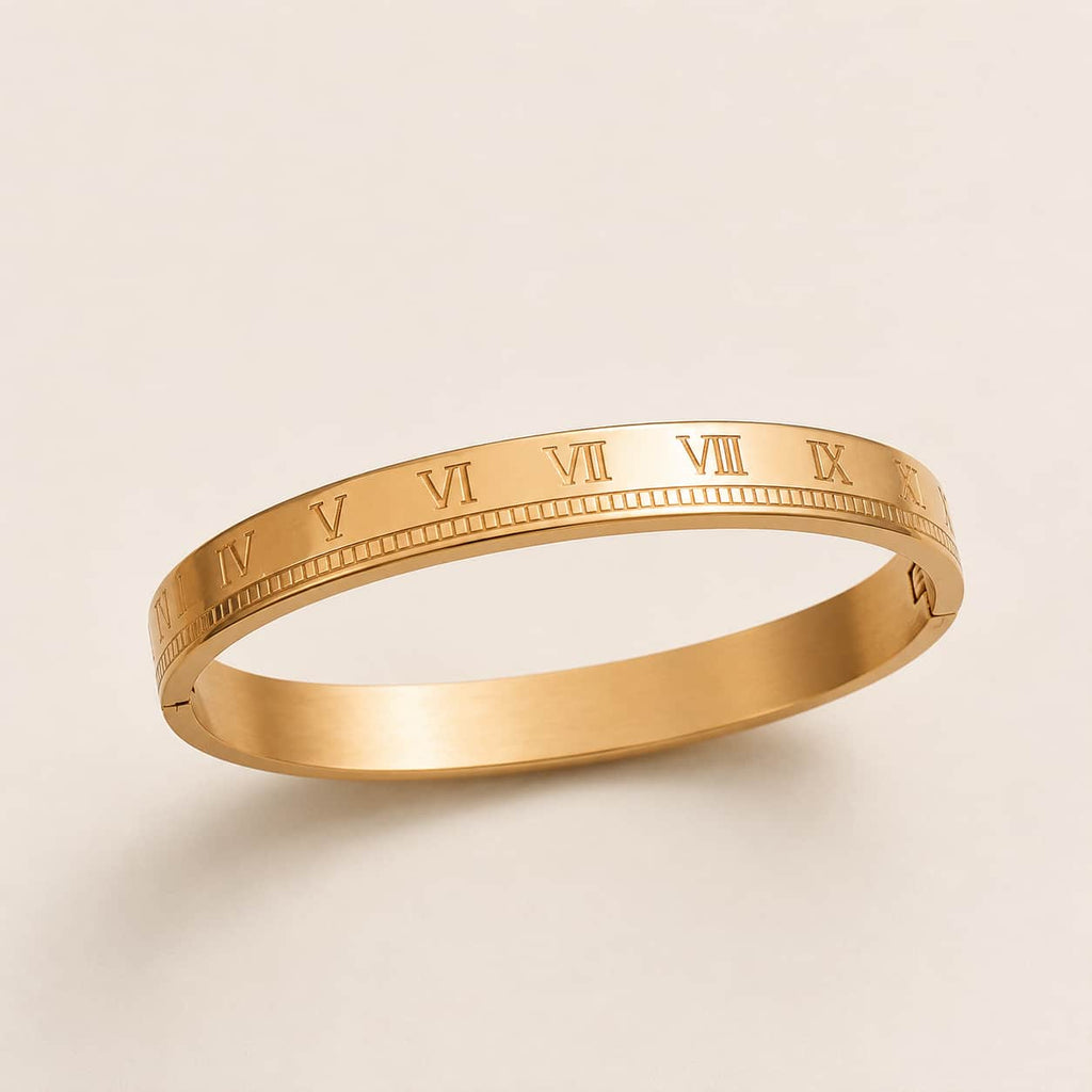 A slim gold bangle bracelet with engraved Roman numerals around the band, shown on a clean, light beige background.