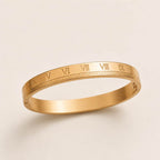 A slim gold bangle bracelet with engraved Roman numerals around the band, shown on a clean, light beige background.