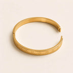 A polished gold-tone cuff bracelet with engraved Roman numerals on the outer band, displayed on a clean, light beige background.