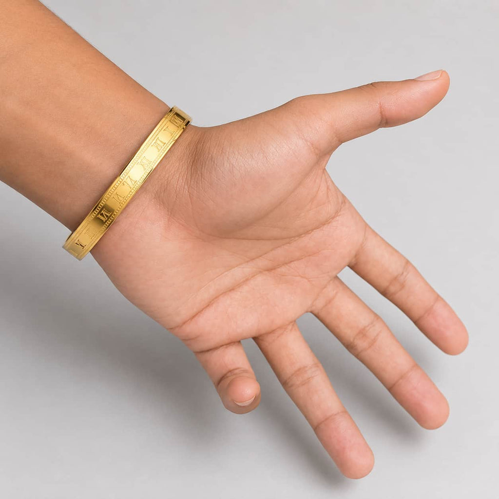 A gold-toned metal bangle bracelet with engraved text, shown worn on a person's wrist with the hand open against a plain light gray background.
