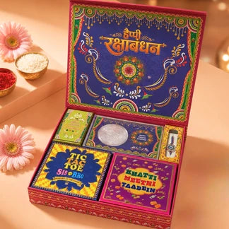 An open decorative gift box for Rakhi Bhandan, shown on a warm beige surface with flowers and small bowls of ingredients around it. The box is brightly colored with a navy blue inner lid, gold and green ornamental details, and several small wrapped items visible inside, including a yellow packet labeled 'Tik Tok Sis Dho' and a purple packet labeled 'Shasti Mantri Taadhin'.